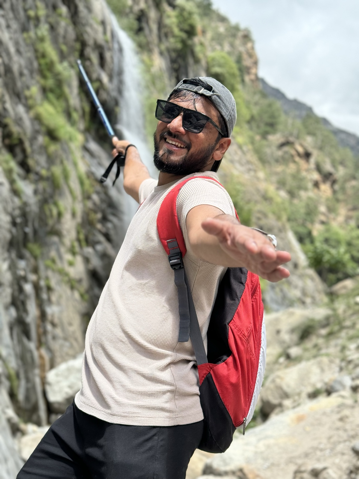 Pratik mid-trek in front of a waterfall with a red backpack, arm flung wide