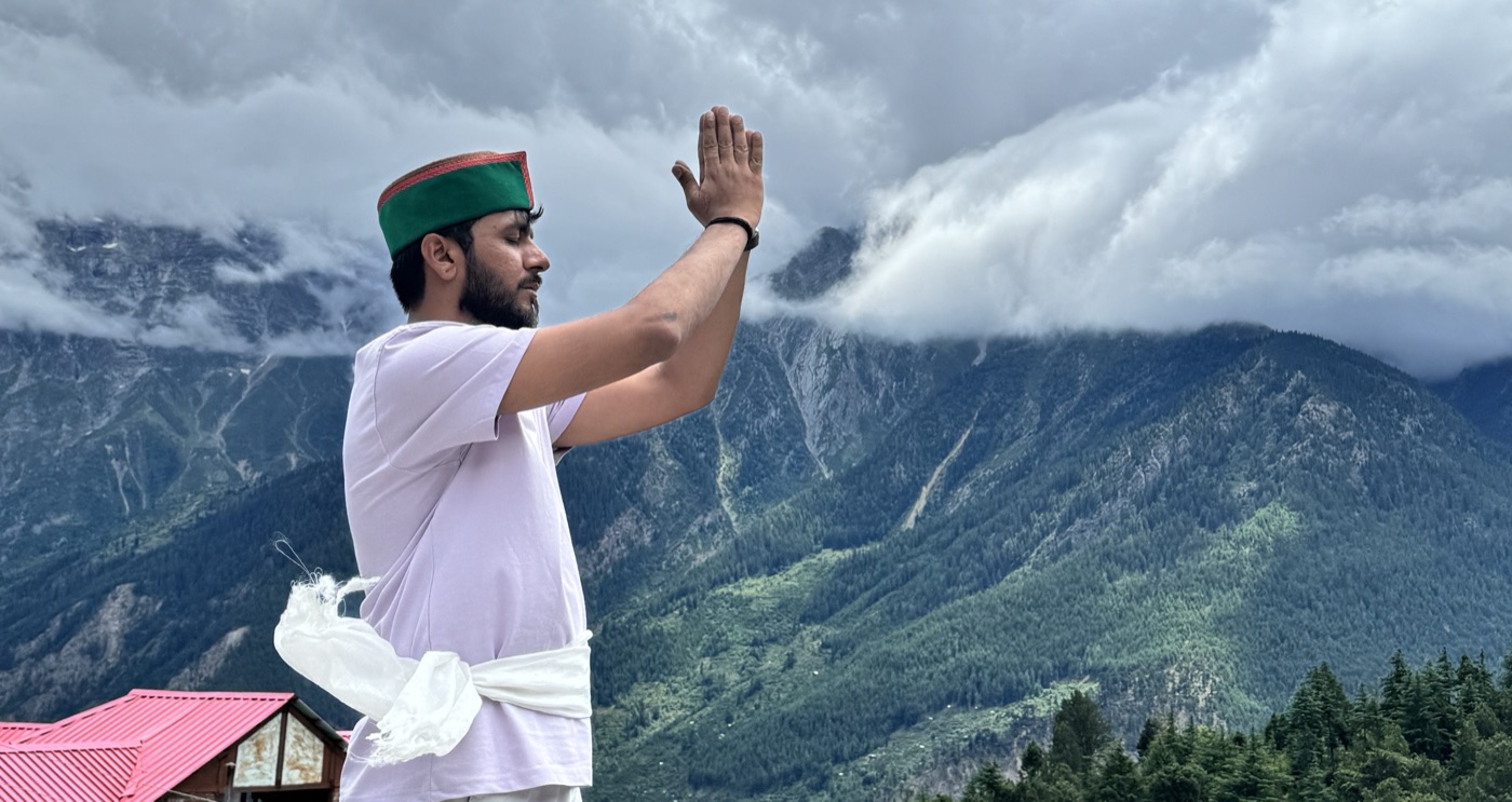 Pratik in a white kurta and Himachali cap, hand raised toward the misty Himalayan mountains
